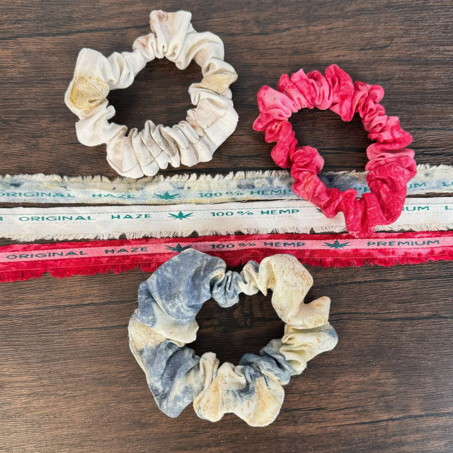 Three hemp eco print scrunchies - eco red, eco blue, eco natural prints. Pictured with three strands of the original fabric scrap edging which states "100% hemp" on the scraps. Background is a dark wooden table.