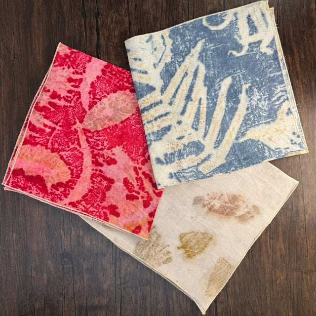 Three eco print hemp bandanas, each folded into a square and stacked partially on top of each other - red eco color is left, blue eco color is top right, and natural eco color is bottom. Pictured on a dark wooden table.