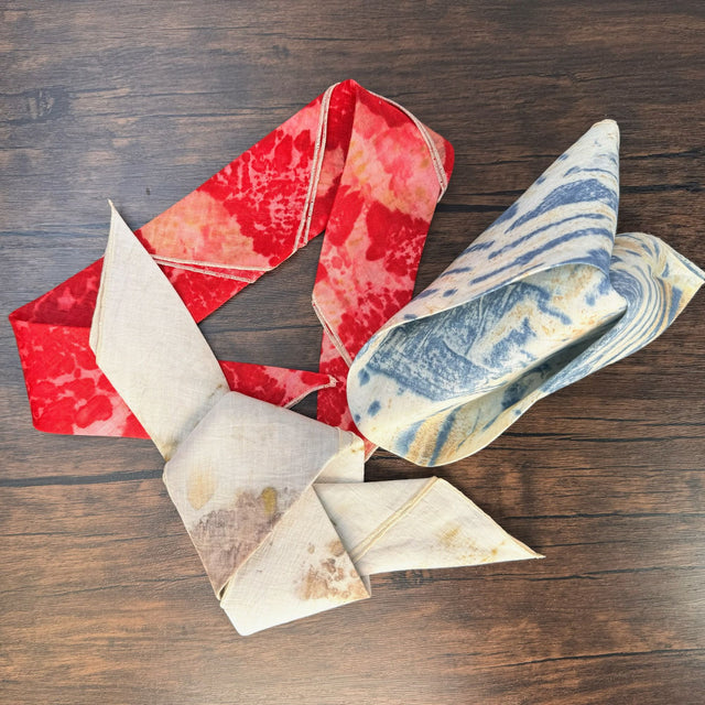 Three eco print hemp bandanas, each folded into and stacked partially on top of each other - red eco color is left and folded like a headband, blue eco color is top right and folded like a triangle party napkin, and natural eco color is bottom and folded like a neck tie. Pictured on a dark wooden table.