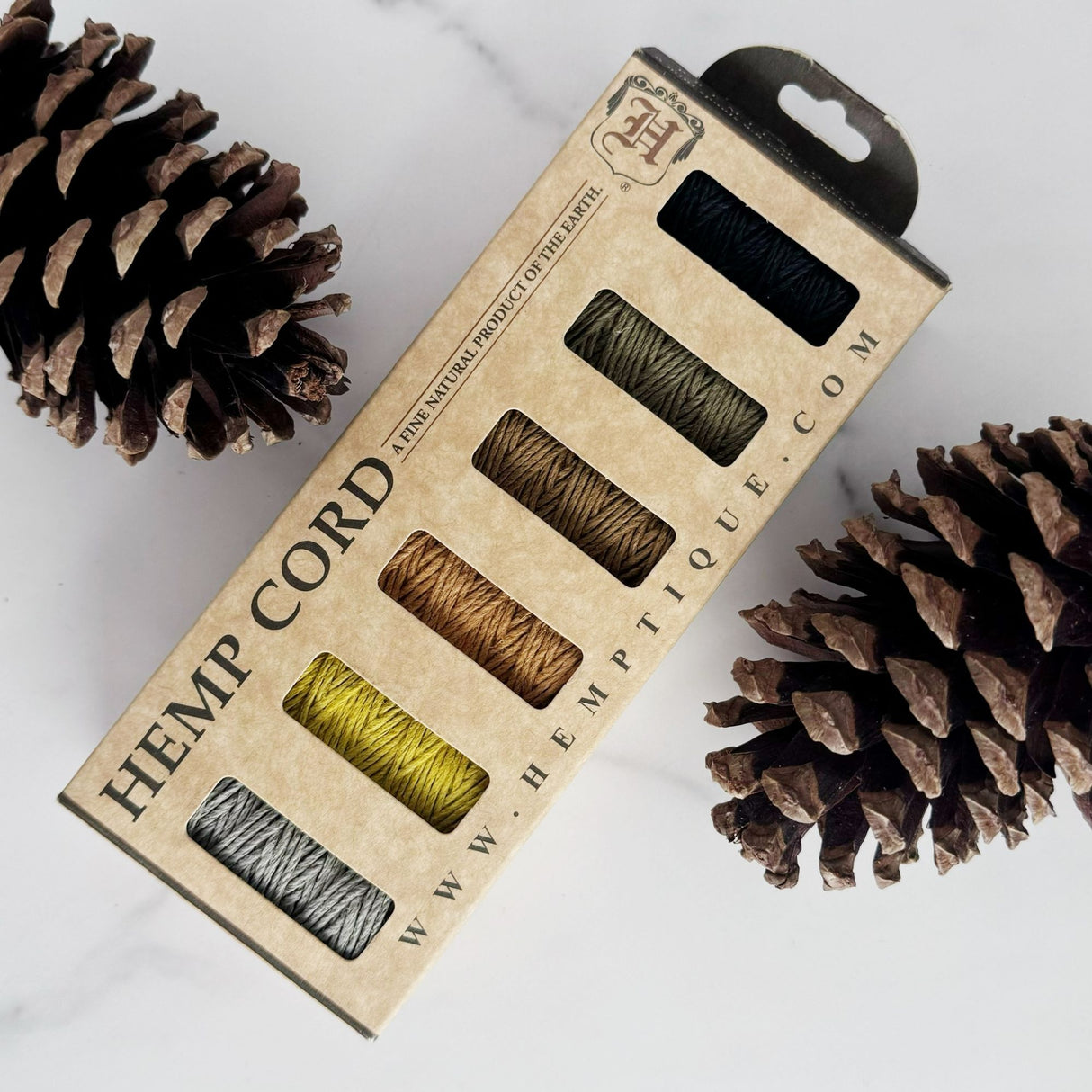 Six rolls of hemp cord - black, grey, sage green, light brown, orange-brown, and yellow. Show in their packaging on a white surface with pinecones in the scene.