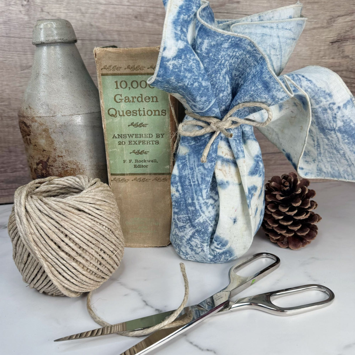 A roll of thick hemp garden cord pictured on a white marble surface with pine cones and antiques in the background. Also shown is a hemp bandana being used as a gift wrap and secured with hemp garden cord tied in a bow.