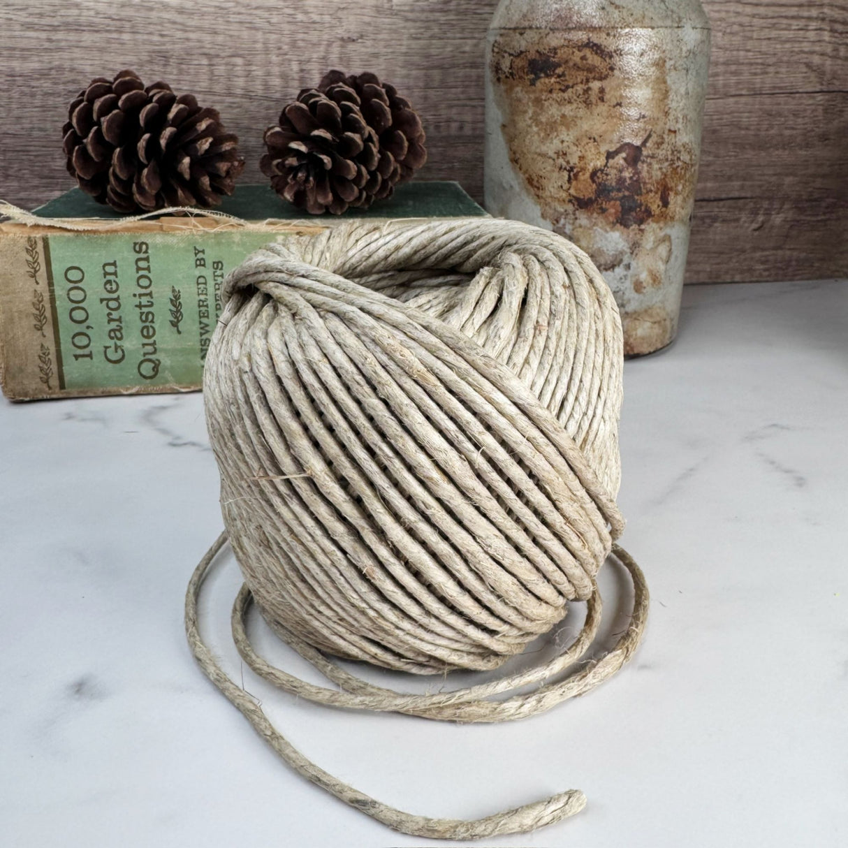 A roll of thick hemp garden cord pictured on a white marble surface with pine cones and antiques in the background.