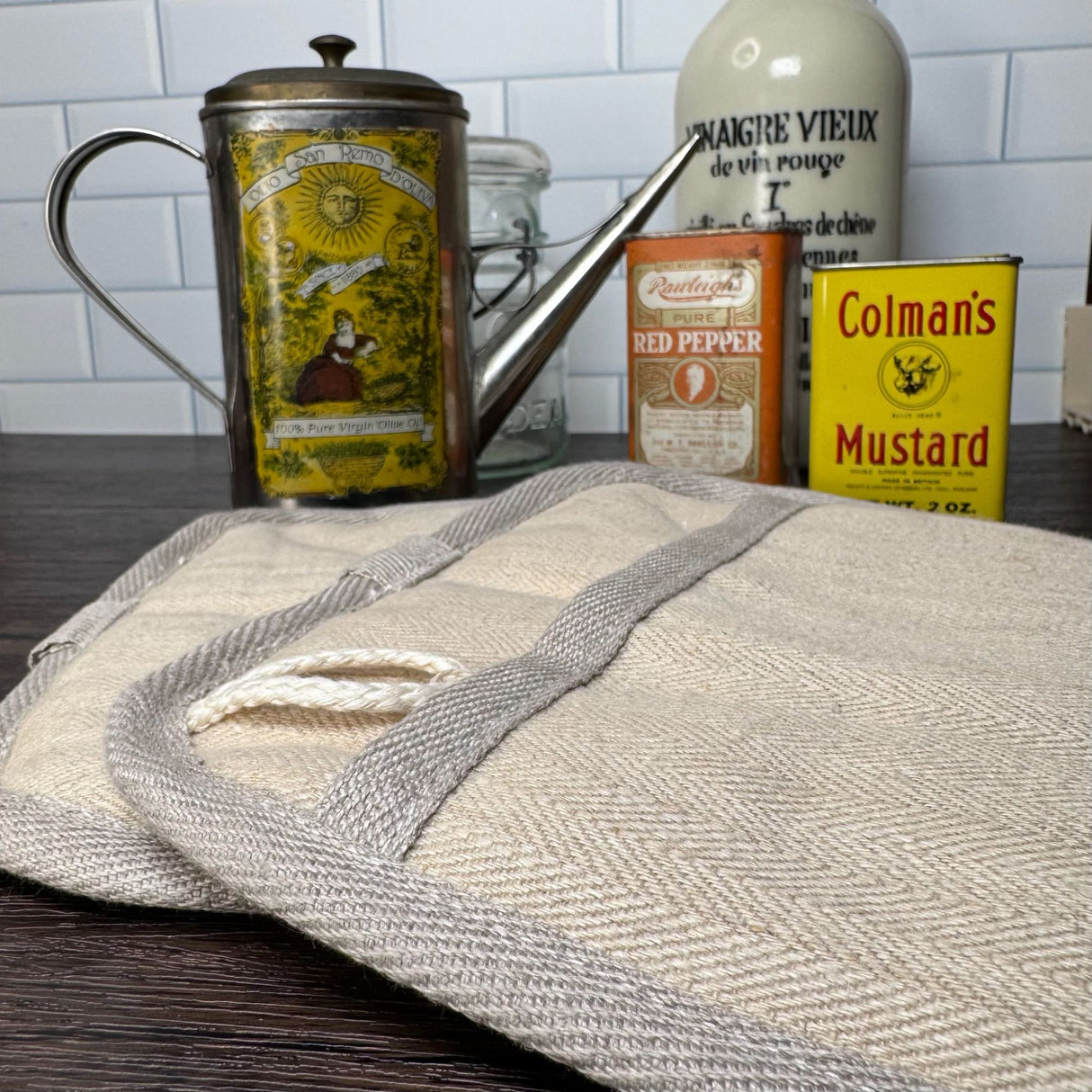 Closeup top/side view of two natural color hemp potholders - background is a variety of antique spice cans. and a mason jar. Pictured on a dark wooden table with a white tile background.