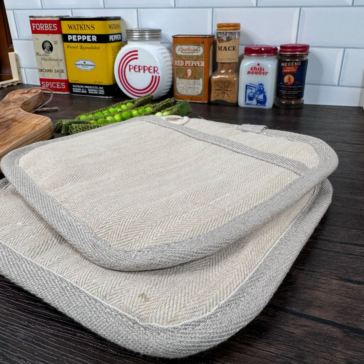 Two natural color hemp potholders partially stacked on top of one another. Ribbing/edging on pot holders is a grey color. Top potholder you can see the loop for hanging. Also pictured with a wooden cutting board, fake snap peas and a variety of antique spice cans and jars in the background.
