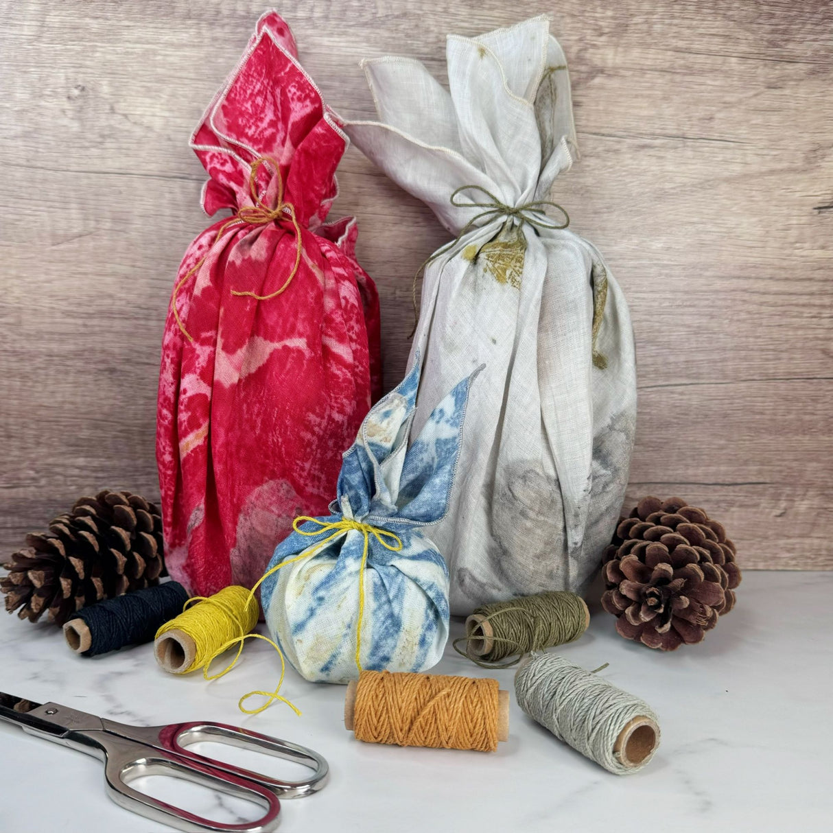 Scene of three items wrapped in hemp bandanas, each secured with a piece of hemp cord making a bow. Five rolls of hemp cord are in the scene (black, yellow, orange, sage green and gray). Also shown with silver scissors. One a white surface with wooden background.