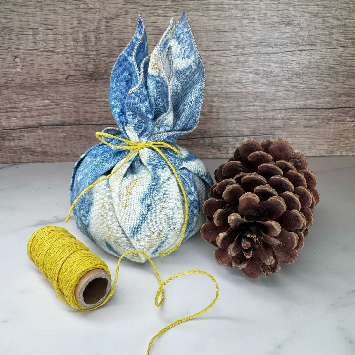 Round object wrapped in a blue hemp napkin and secured with a piece of yellow hemp cord tied in a bow. Also pictured with a spool of yellow hemp cord and a pinecone. One a white surface with a wooden background.