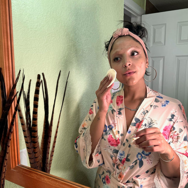Model wearing a floral lightweight bathrobe is using a hemp facial round to clean her face while looking in the mirror. Model is also wearing large hoop earrings and a pink headband.