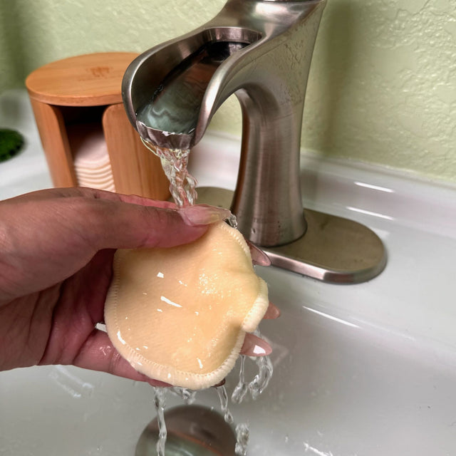 Woman's hands holding a single hemp facial round under running water. In the background the full bamboo box with a stack of facial rounds can be seen.