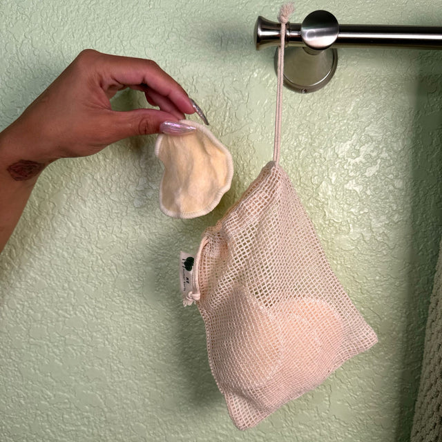 A hand is dropping a used hemp facial round into the mesh washing bag which is hanging on a towel rack.