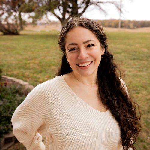 Profile picture for Kristen Williams. Wearing a white sweater, hair half up and green field in the background