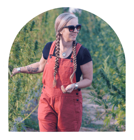 Picture of Lauren Gibbs on a hemp farm, wearing red hemp overalls and a black tee shirt and touching the hemp plants.