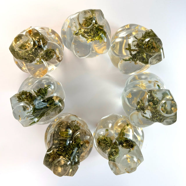 Top view of seven hand poured resin figurines in a circle - all are of a full bodied woman, with hemp buds at heart-center and gold flake throughout to show the variation in the art. Scene is on a white surface with a white background.