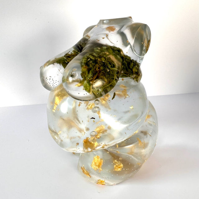 Closeup view of a hand poured resin figurine of a full bodied woman, with hemp buds at heart-center and gold flake throughout. Figurine is sitting on a white surface with a white background.