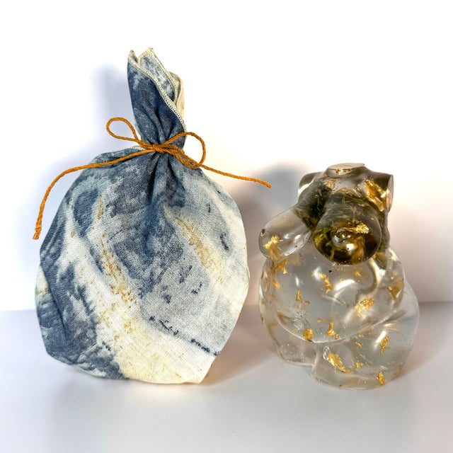 Side by side view of two hand poured resin figurine of a full bodied woman, with hemp buds at heart-center and gold flake throughout to show the variation in the art. Left figurine is wrapped in hemp material gift wrap ties with a hemp cord bow.
Scene is on a white surface with a white background.