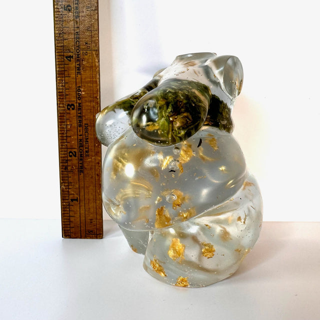 Closeup view of one hand poured resin figurine of a full bodied woman, with hemp buds at heart-center and gold flake throughout to show the variation in the art. Figurine is picture next to a ruler to show the size (4 inches tall). Scene is on a white surface with a white background.