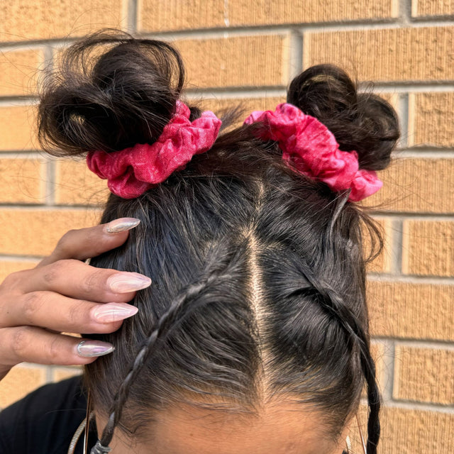 Two red eco print hemp scrunchies securing pig-tail buns of dark hair on a model. Model's hands with long nails touching her head, background is a yellow-ish brick wall.