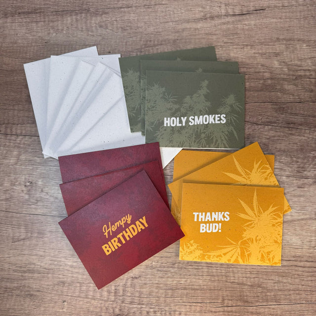 3 red hemp paper greeting cards with the words "hempy birthday." 3 - green greeting cards with the words "holy smokes." 3 - yellow greeting cards with the words "thanks bud." Pictured with nine hemp paper envelopes all on a dark wooden background.