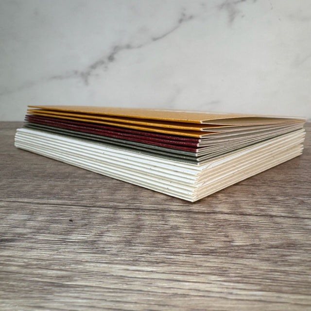 Side view of a stack of 9 greeting cards (3 green + 3 red + 3 yellow) on top of nine envelopes that are off-white color. Pictured on a dark wood table with a white marble background.