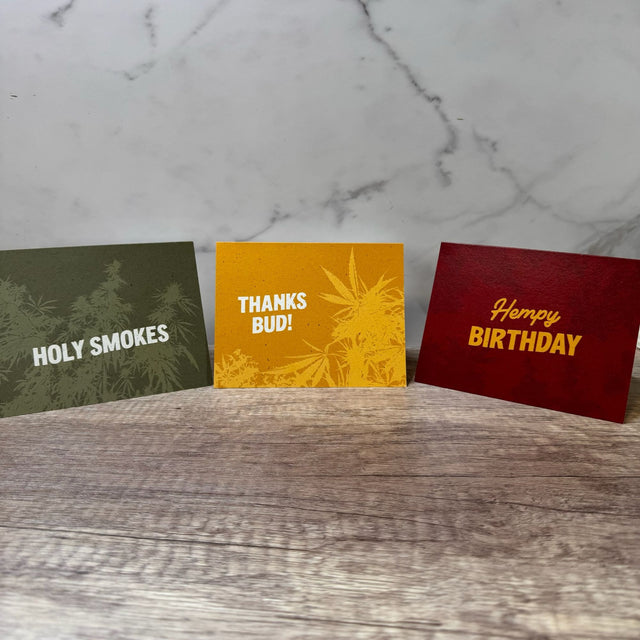 Three hemp paper greeting cards folded to stand upright - left to right: green card with words "holy smokes", yellow car with words "thanks bud" and red card with words "hempy birthday". All cards have an underlying graphic of hemp plants. Pictured on a dark wooden table with a white marble background.