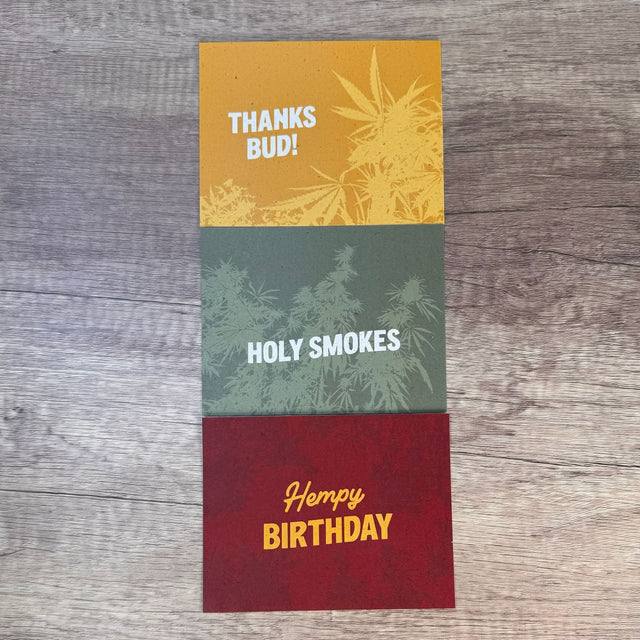Three hemp paper greeting cards folded to lay flat on a table - top to bottom: yellow car with words "thanks bud", green card with words "holy smokes", and red card with words "hempy birthday". All cards have an underlying graphic of hemp plants. Pictured on a dark wooden table.