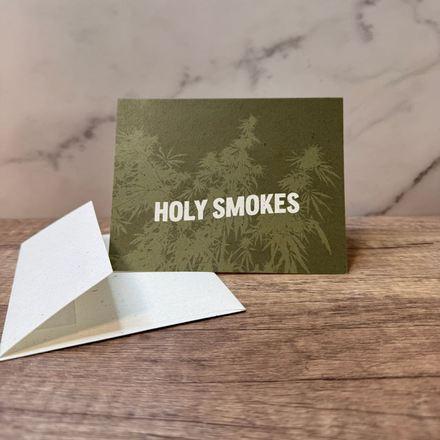 Green hemp paper greeting card with white words that read "holy smokes." Graphic on front of card includes hemp plant underlying the words and in light green. Pictured with an unsealed hemp paper envelope.