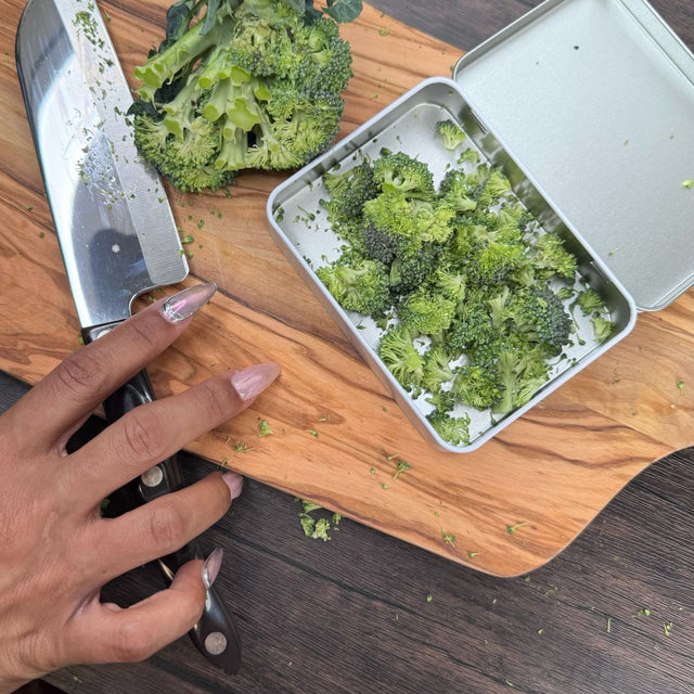Open view of the pocket tin - inside is silver. Tin is filled with chopping broccoli. Also in the picture are a wooden cutting board, knife, and hand with long fingernails.