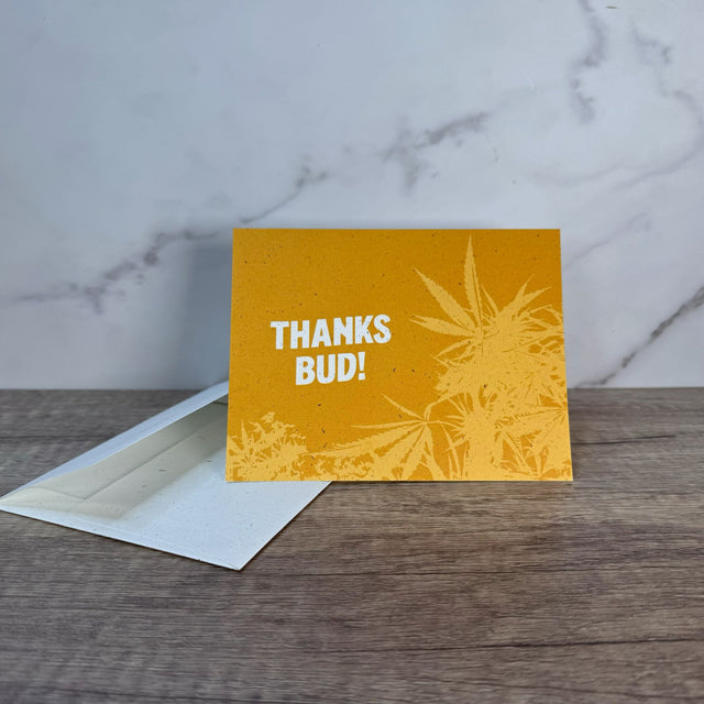 Yellow hemp paper greeting card with white words that read "Thanks Bud!" Graphic on front of card includes hemp plant underlying the words and in yellow green. Pictured with an unsealed hemp paper envelope.