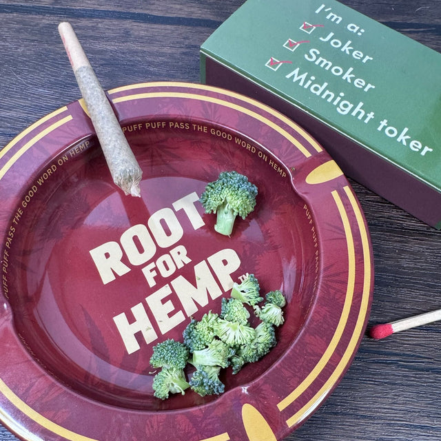 Round metal ash tray with red and gold with dark red leaf pattern, "Root for Hemp" on center of dish, "puff puff pass the good word on Hemp" on the inside rim. Ash tray has broccoli inside and a joint sitting on the rim. Pictured with a book of matches.