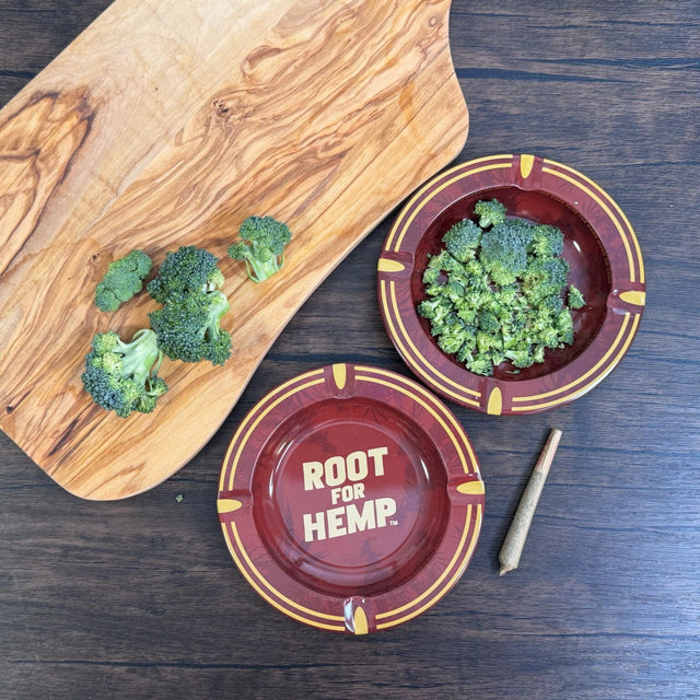 Top own view of two red and gold round metal ash trays Red and gold with dark red leaf pattern, "Root for Hemp" on center of dish. Pictured with a wooden cutting board, chopped broccoli and a joint.
