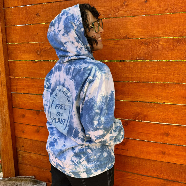 Side/back view of a model wearing an indigo tie dye hoodie with a the words "free the people, free the plant" screen-printed on the back. Model has the hood over her head, and the side of her face can been seen - she has dark hair and green glasses. The background of the scene is a wooden fence.