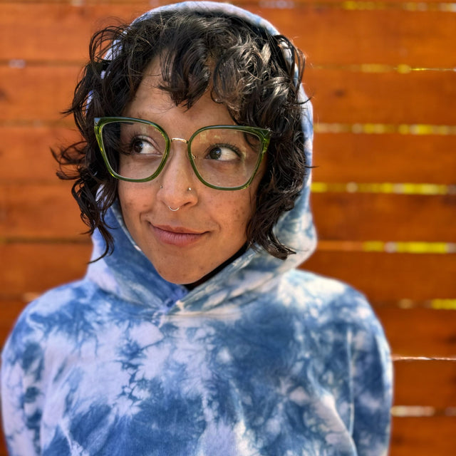 Closeup headshot view of a model wearing an indigo tie-dyed hoodie. She has the hood over her head and her dark hair can be seen coming out of the front. She is also wearing green glasses and gazing up away from the camera angle