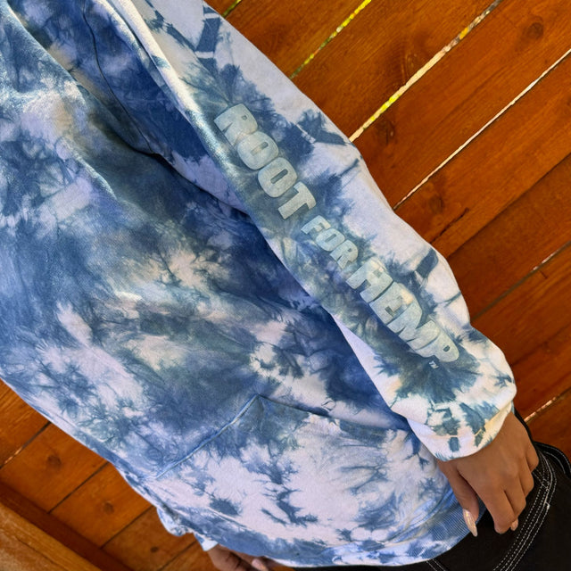Left arm view of a model wearing an Indigo tie dye hoodie with a the words "root for hemp" printed down the arm in bold white lettering.