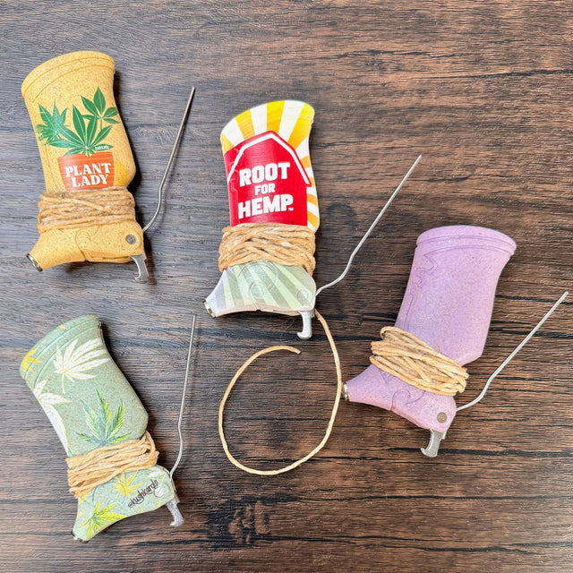 Four toker pokers pictures on a wooden surface. Each has a wrapping of hemp wick around the bottom. Four different toker designs are in the photo - top left is a yellow toker with a hemp plant and the words "plant lady", bottom left is a green toker with green, white and yellow hemp leaves, middle is a toker with the words root for hemp in white on a red barn and sunburst in the background, and the right toker is a plum color with no images.