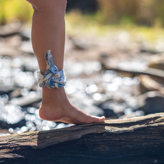 A blue hemp bandana is wrapped around a person's ankle. Foot is bare foot and standing on a log. A river can been seen in the background.