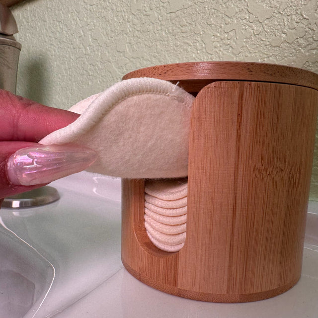 Cylindrical Bamboo box with hemp facial rounds stacked inside. Box has a slit opening down the side for dispensing. Pictured with a woman wearing long tails pulling one round out of the box.
