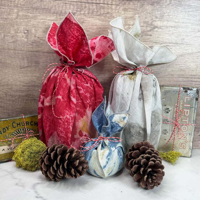 Hemp bandana being used as gift wrap on two items that are each tall and cylindrical (one wrapped in red eco print, the other wrapped in natural eco print). Another smaller item is wrapped in a blue eco print napkin. Gift wraps are tied with red and white hemp cord. Also pictured are two pinecones, moss, and two antique cans.