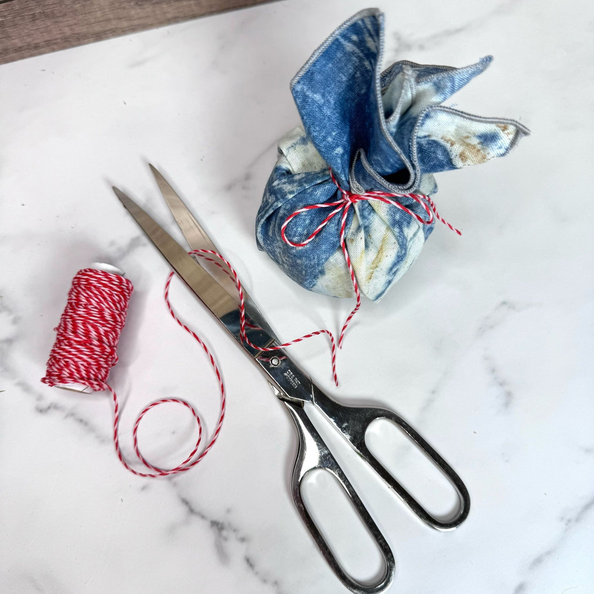 Red and white spiral hemp cord partially unwound on a white marble table. Pictured with classic silver colored scissors and a small object wraps like a present using the blue eco print napkin and red and white hemp cord to secure.