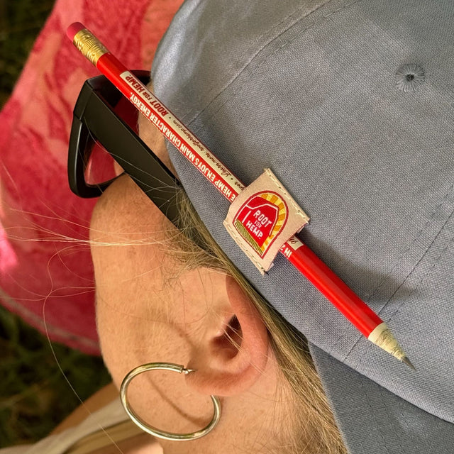 Closeup view of the pencil holder on the blue root for hemp vote hat in use. Pencil is red with hemp educational messaging. The hat is on a model with blond hair, wearing black glasses and silver hoop earrings.