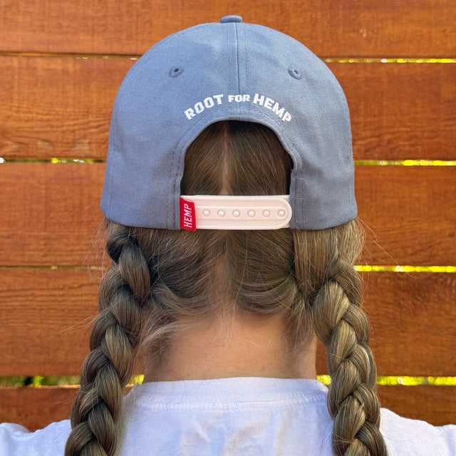 Back view of the hemp vote hat on a model. Back of hat is blue with the words root for hemp above the snap-back and a red hemp tag on the snapback. Model has dirty blonde pigtail braids.