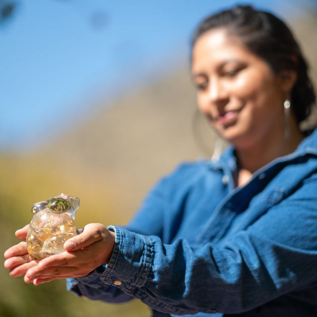 Woman wearing a blue denim shirt holding her hands out in front of her with a hand poured resin figurine of a full bodied woman, with hemp buds at heart-center and gold flake throughout.