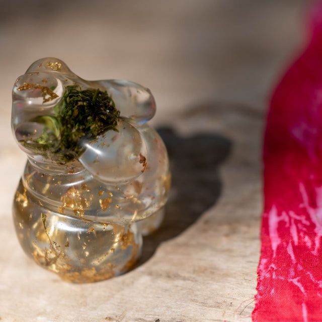 Closeup side view of a hand poured resin figurine of a full bodied woman, with hemp buds at heart-center and gold flake throughout. Figurine is sitting on a wooden surface. A piece of red hemp fabric can be seen in the right side of the image.
