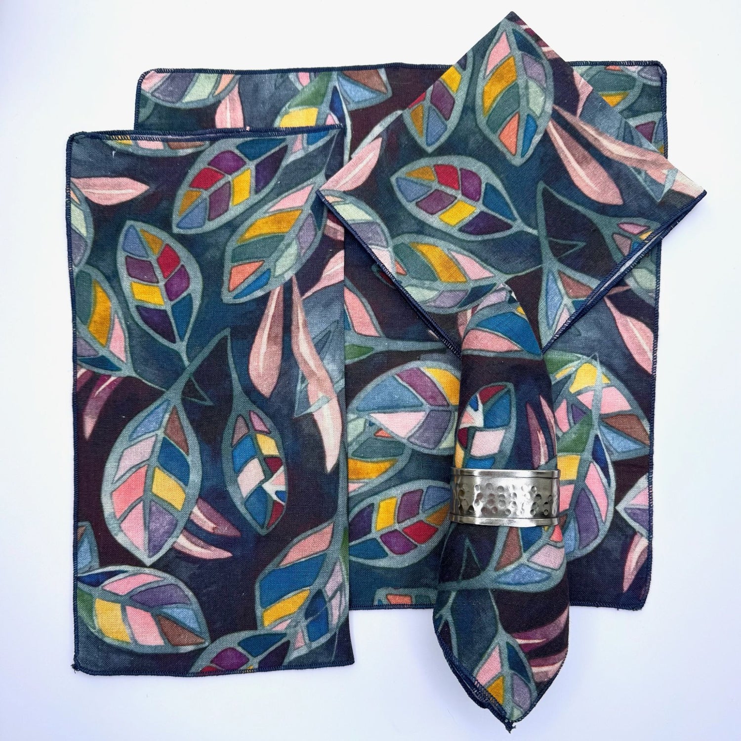 100% hemp table napkin with a multi color flower print with purple/blue background, pictured with 5 different ways to fold the napkin.  Napkins have black threading on the edge.