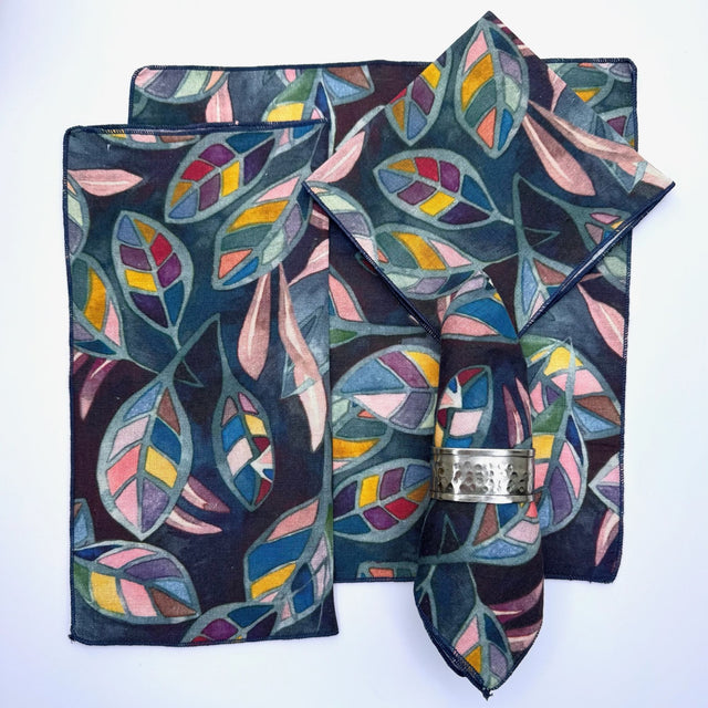 100% hemp table napkin with a multi color flower print with purple/blue background, pictured with 5 different ways to fold the napkin.  Napkins have black threading on the edge.