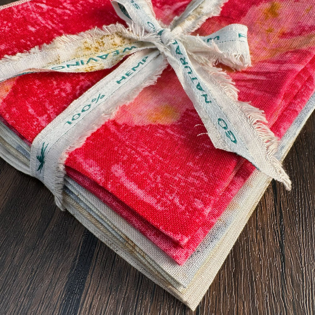 Closeup view of 100% Hemp table napkins, 12 napkins folded into squares and stacked. Stack is secured with a scrap of hemp fabric tied in a bow, on a wooden table background. Bottom of stack is natural color napkins, middle of stack is blue color napkins, and top of stack is red napkins.