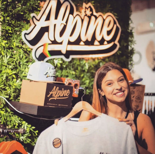 Girl holding a white sweatshirt with alpine hat logo on large sign in the background.