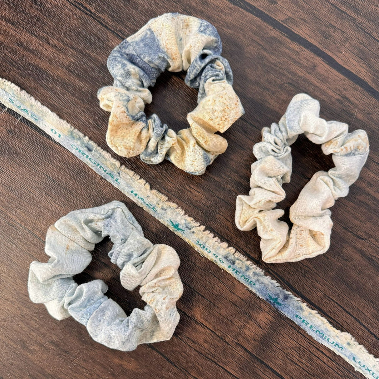 Three blue eco print hemp scrunchies, pictured on a dark wooden table and pictured with a piece of hemp fabric scrap of the same fabric that reads "100% hemp"