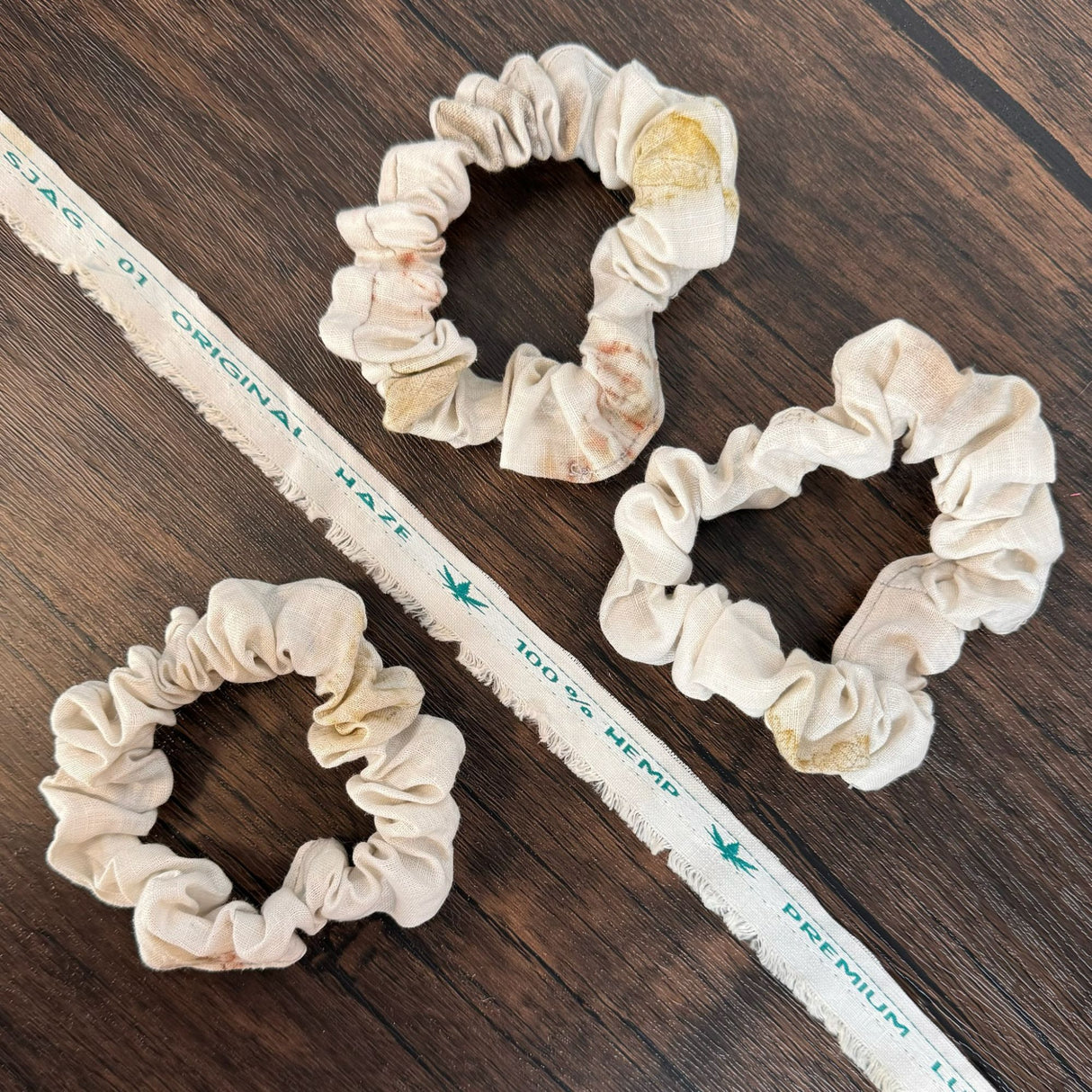 Three natrual eco print hemp scrunchies, pictured on a dark wooden table and pictured with a piece of hemp fabric scrap of the same fabric that reads "100% hemp"