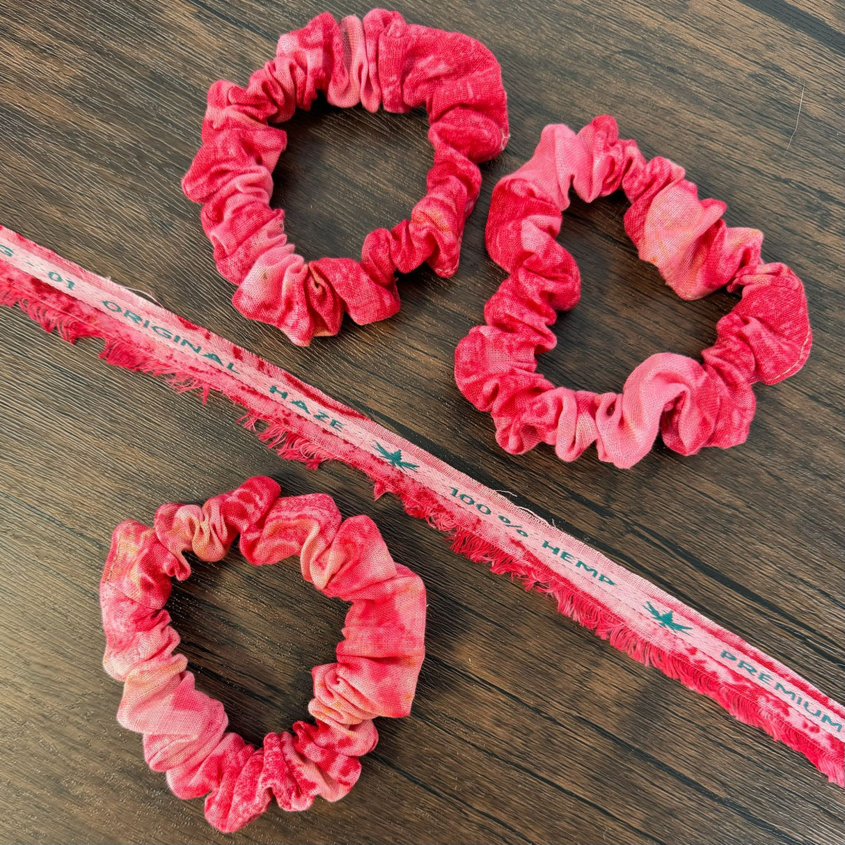 Three red eco print hemp scrunchies, pictured on a dark wooden table and pictured with a piece of hemp fabric scrap of the same fabric that reads "100% hemp"