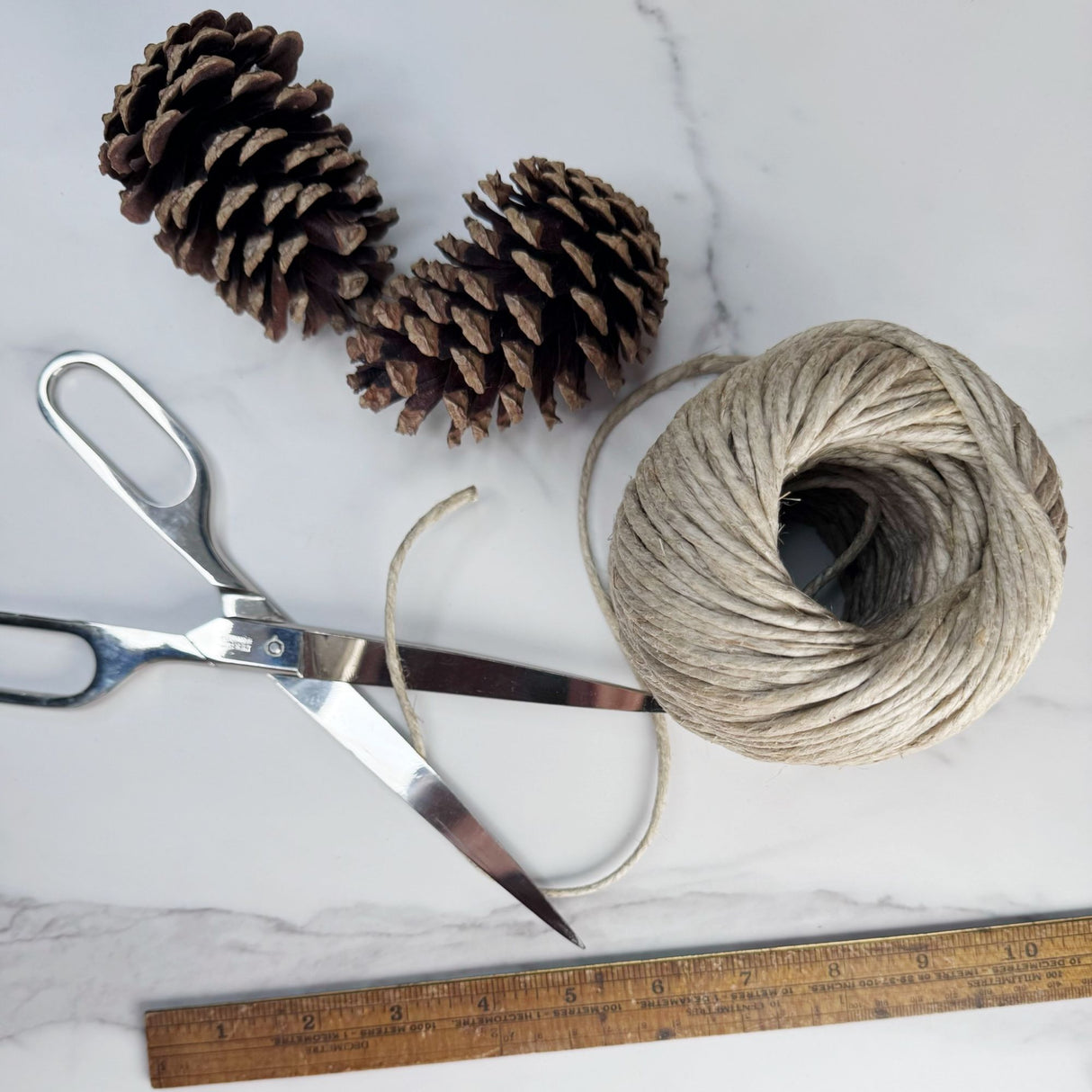 Top view of a roll of thick hemp garden cord pictured on a white marble surface with pine cones and silver scissors next to it.  Also show with a ruler to show the size of the cord roll.  