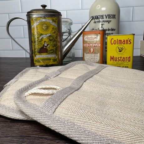 Closeup top/side view of two natural color hemp potholders - background is a variety of antique spice cans. and a mason jar.  Pictured on a dark wooden table with a white tile background.